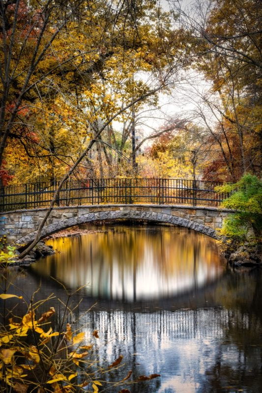 A stone footbridge over a still pond amidst yellow leaves in autumn.