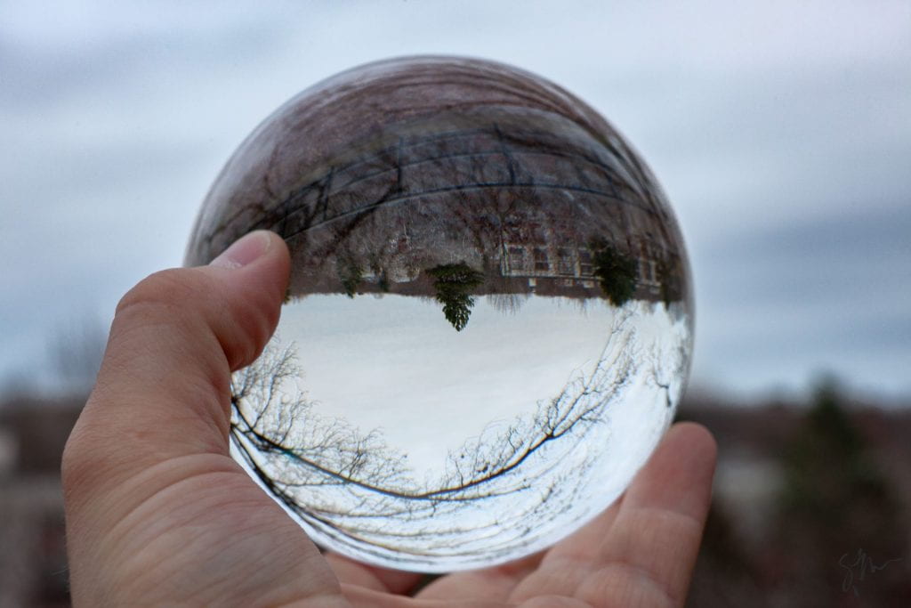 A hand holding a crystal ball that is in crisp focus and reflects the surroundings upside down. A Pine tree in the center of the inverted image points down, building hang from the ground which is at the top of the crystal ball. Outside the crystal ball, the surrounding are situated as we experience them, with the sky above and the ground at the bottom... though that is all out of focus.