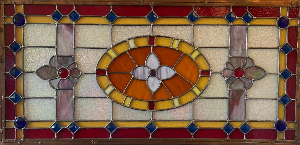 A stained glass mosaic geometric pattern - red rectangle and yellow rectangles interspered with blue squares make the frame with 2 symmetric fleur de lis and a white flower in an orange oval.