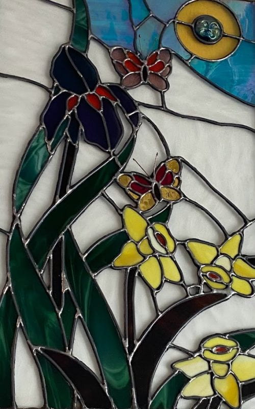 A stained glass mosaic with three yellow daffodils in the bottom right corner, a purple iris spanning the left side of the piece, and two butterflies.