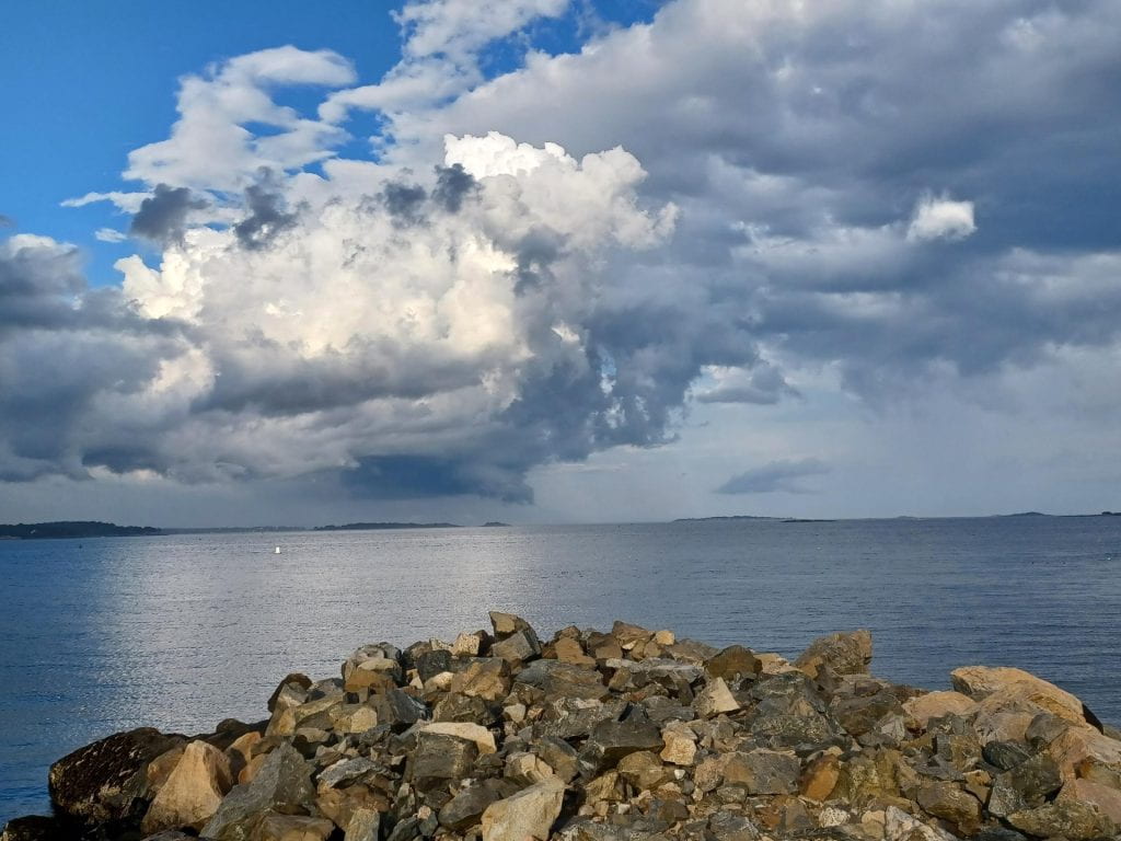 A seascape. In the foreground a rocky beach; the sea behind it is calm, but there is a massive stormcloud rolling in far in the distance, with distant curtains of rain. To the top left there is a corner of blue sky.