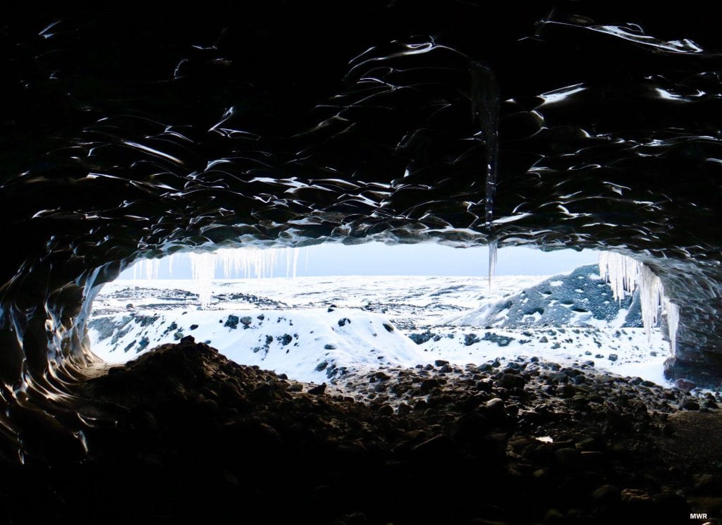 A blue ice cave with icicles coming down from the opening and snow and rocks on the ground.