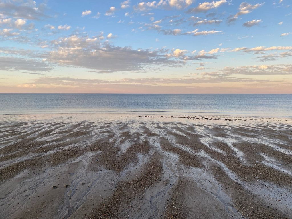 Wet sand patches form a pattern of rivulets which end on a serene light blue sea that expands till the horizon. The sky above is orange pink and then blue with scattered clouds, and its colors reflect back to the beach and the water.