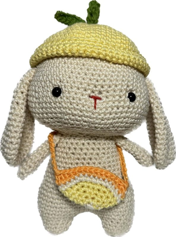 A very cute cream-colored crocheted bunny with a little yellow hat with leaves on top and an orange-and-white striped shoulder bag.