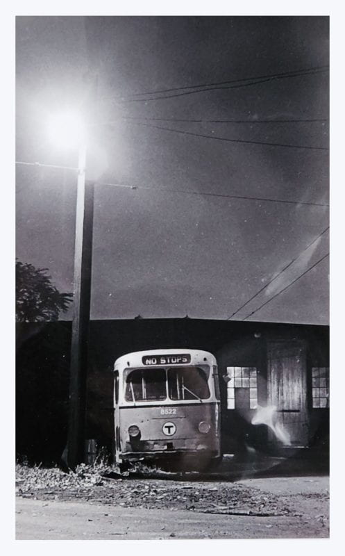 A black and white photograph of a 1960s MBTA bus, parked at the Eliot Shops bus yard (now the JFK School of Government). The destination reads NO STOPS, and the streetlight above illuminates the dusty road below.