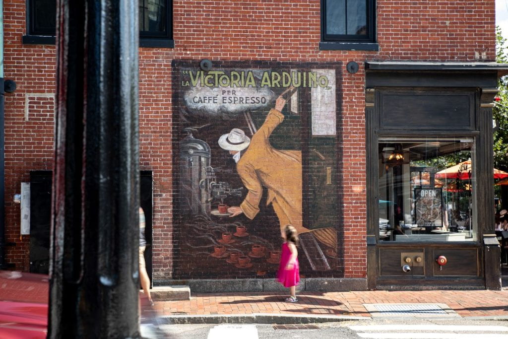 A young girl in a bright pink dress is blurred as she runs on a cobbled sidewalk past a painted wall mural on a brick wall. The wall mural features a vintage Caffe Espresso ad.