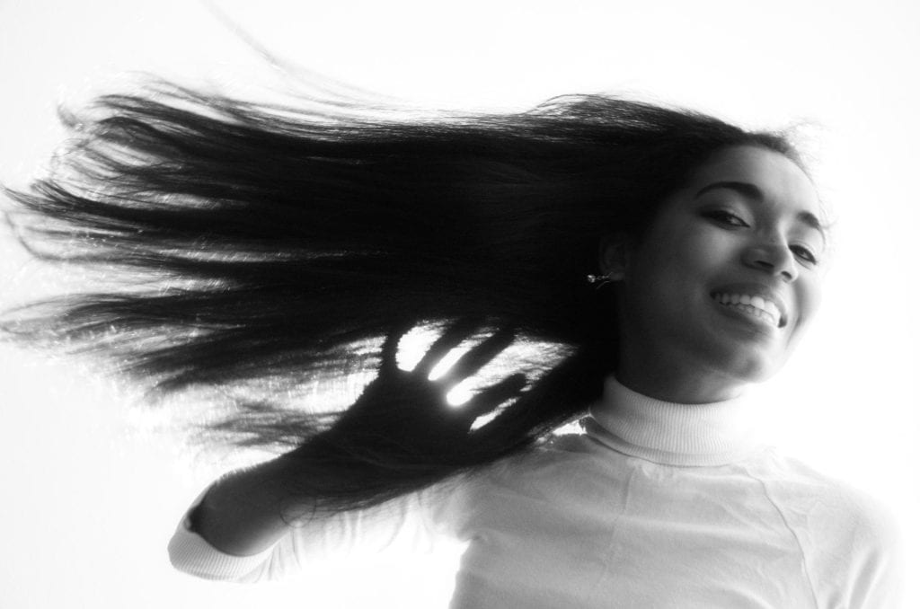 A young woman faces downward towards the camera. There is a big smile on her face and her long hair is swept away by the wind, floating like a banner. The photo is in black and white.