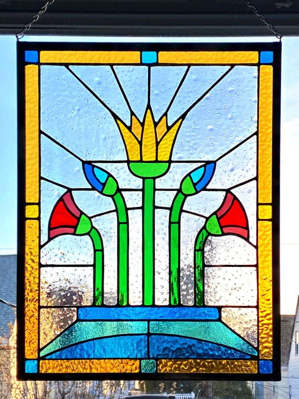 Part of a window hosts a rectangle stained glass with six colorful flower buds of three different kinds of plants. The rectangle is framed by a yellow line with scarce light blue and golden squares that are also glass-stained.