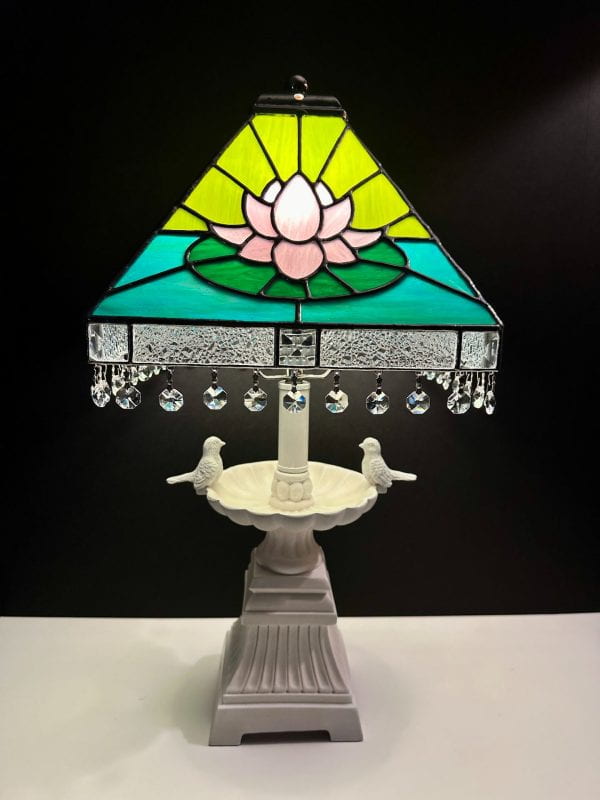 A table lamp whose base is white and reminds a fountain with two pigeons, one on each side. The lamp’s hat is made of stained glass, brightly colored with light green, green, and azure, and with a baby pink water lily flower in the middle. Clear hexagonal little crystals are hanging perimetrically at the bottom edge of the hat.