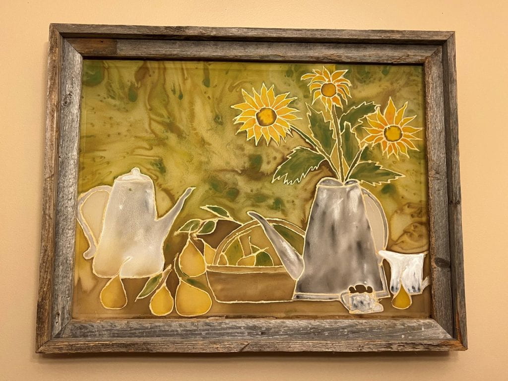 A painting on silk. A still life, with a silver coffeepot and silver teapot/flowerpot holding daisies. Pears lie on the table and in a basket. The background is a swirl of gold and green.