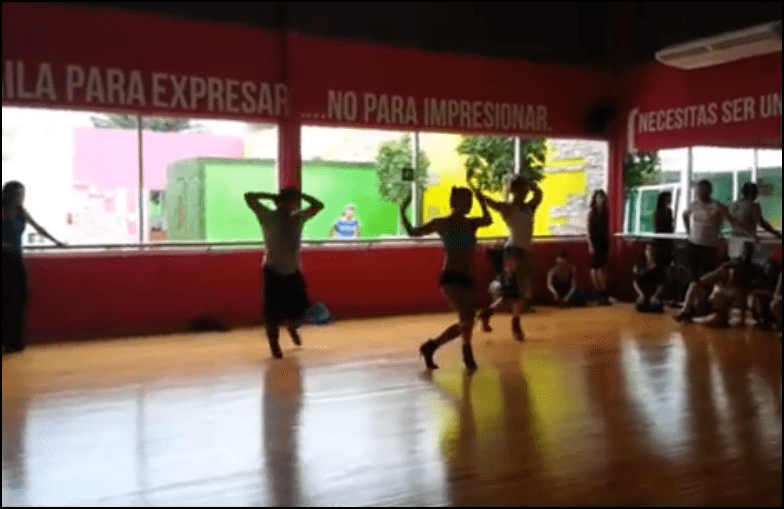 Screenshot from a video of the dance class. A number of dancers in a studio follow the lead of the instructor.