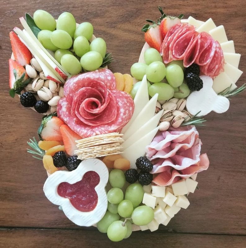 Grapes, slices of strawberries and apricots, berries, shelled pistachios, white and yellow cheese cuts, cured meats, some crackers, and some jam placed together colorfully to form the layout of Mickey Mouse's head.