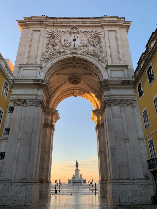 A monumental 100 foot tall gray stone archway topped with a clock. The sunrise is coming up behind the archway, casting a light onto the plaza and statue behind it.