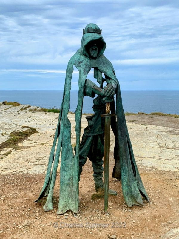 A photo of a weathered bronze statue on a desolate hilltop with the ocean and sky in the background. The statue is of a cloaked male figure, wearing a crown and holding a sword pointing toward the ground beneath him. The figure is hooded and his face can't be seen. The statue is designed so that large portions of it are missing, creating negative space which the surrounding landscape can be seen through, giving the figure a ghostly appearance.