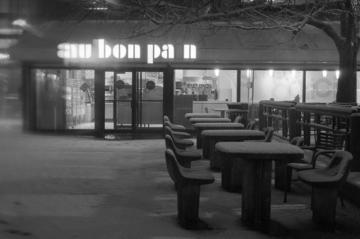 An outdoor view in black and white of nighttime, snow, an illuminated “au bon pain” cafe sign with the light of the letter “i” turned off, and steamy windows blurring those inside.